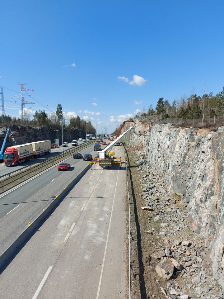 I Vanda på Ring III (stamväg 50) vid Kalkkikallio utförs bergrensning på bergskärningarna bredvid vägen 21.4.2021. Den orangefärgade grävmaskinen i bakgrunden gör en maskinell bergrensning och från personkorgen i förgrunden görs bergrensning för hand.