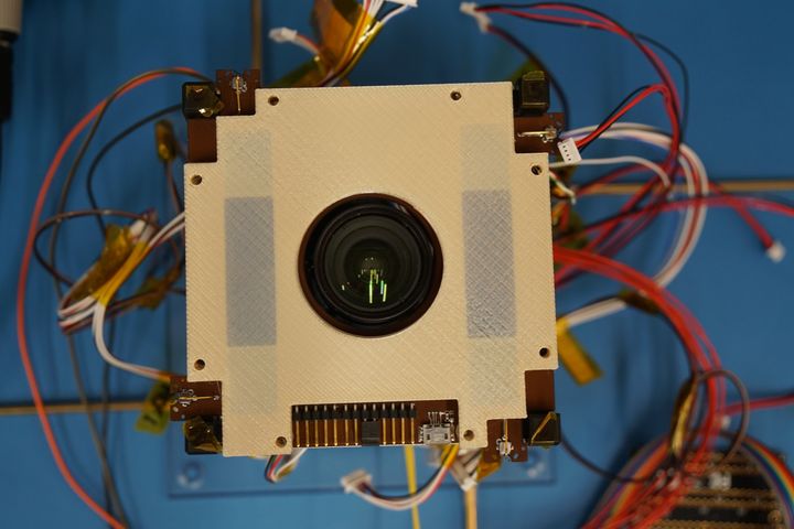 Rakenteilla oleva Suomi 100 -satelliitti suoraan ylhäältä katsottuna. Vaalea osa on eräs satelliitin erikoisuuksista: Suomessa kehitetyllä tekniikalla 3D-tulostettu avaruuskelpoinen osa, jonka sisällä on avaruussäätä havaitsevan radioinstrumentin antenni. Keskellä näkyy satelliitin kamera. Kuva otettu elokuussa. Kuva: Jari Mäkinen