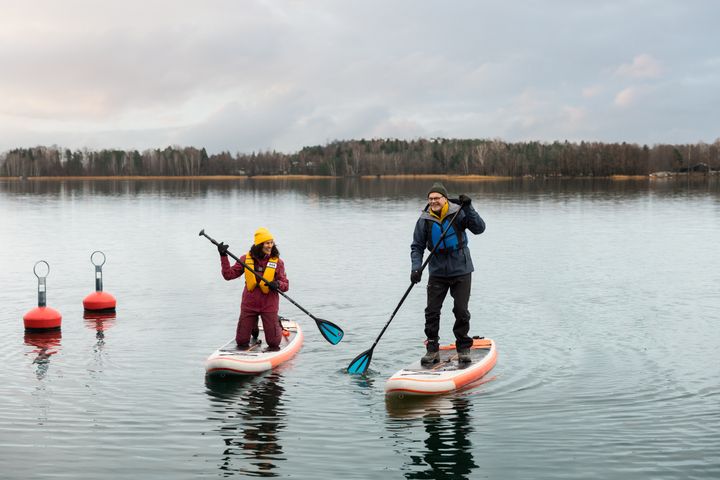 Kaksi henkilöä meloo SUP-laudoilla syksyisellä järvellä. Taustalla metsää.