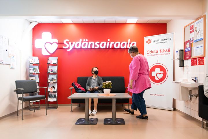 Odotustilassa sairaalassa yksi henkilö istuu sohvalla ja toinen henkilö lähestyy istujaa, kutsuen hänet vastaanotolle.