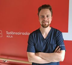 Henkilökuvassa hymyilevä erikoistuva lääkäri Jani Rankinen on pukeutunut sairaalan siniseen työasuun, taustalla punainen seinä ja teksti "Sydänsairaala"