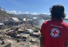 Person i röd Röda Korset-tröja ser ut över förödelsen efter en tyfon i Filippinerna. Hus är förstörda och naturen är skadad.