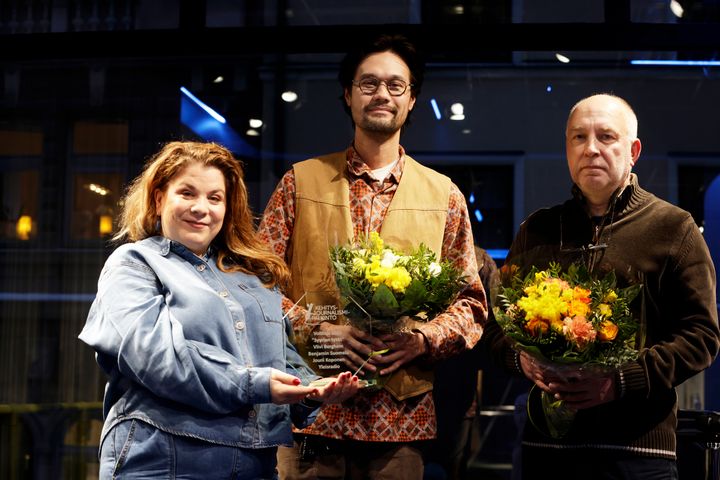 Tre personer står på rad och håller i en vinnarpokal samt blomsterbuketter.