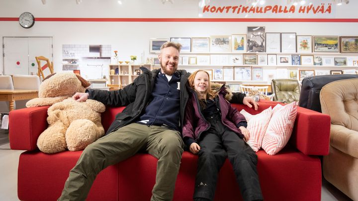 Kuvassa mies ja tyttö istuvat punaisella sohvalle Punaisen Ristin Kontti secondhand-ketjun myymälässä
