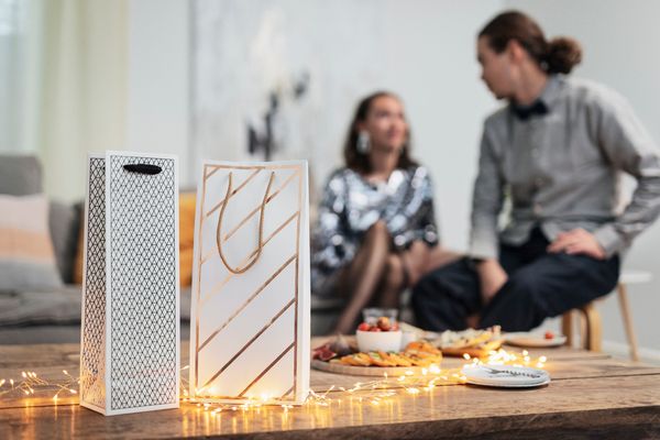 Kaksi henkilöä istuu taka-alalla, juhlallisesti pukeutuneina. Edustalla on lahjakasseja ja pöydällä on tarjolla naposteltavia. Jouluvalot valaisevat pöytää.