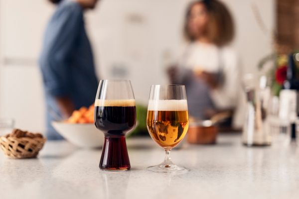 Kaksi lasia olutta keittiön pöydällä, taustalla ihmisiä keskustelemassa.