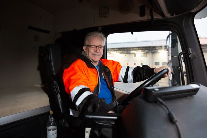 Kuljettaja istuu kuorma-auton ohjaamossa oranssi turvatakki päällään.