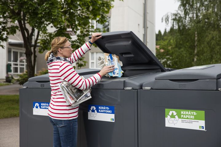 Nainen laittaa pahvia kartonkikeräykseen. Kainalossa on lehtiä seuraavaksi paperinkeräykseen laitettavaksi.