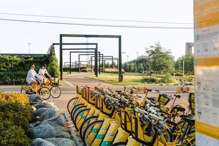 Kaksi ihmistä ajavat keltaisilla kaupunkipyörillä puistossa. Etualalla on rivi pysäköityjä kaupunkipyöriä.