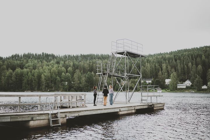Valokuvassa kolme lasta seisoo suomalaisessa järvimaisemassa laiturin päässä ja keskustelee keskenään. Järven vastarannalla näkyy metsää.