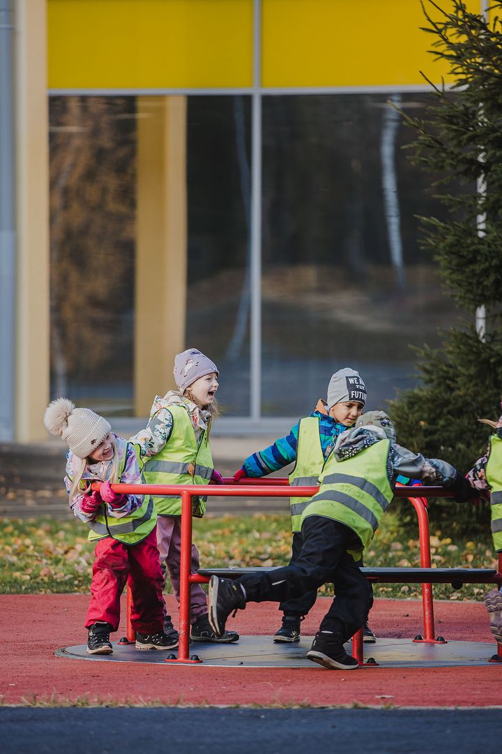 Lapset leikkivät ulkona heijastinliivit päällä. Tunnelma on iloinen ja vauhdikas.