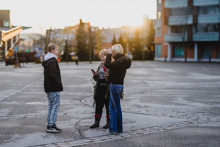 Kolme nuorta keskustelee ulkona kaupunkiympäristössä. Taustalla näkyy rakennuksia ja aurinko laskee.