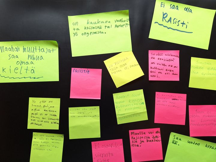 Värikkäitä post-it-lappuja seinällä, joissa on lasten kirjoittamia viestejä maahanmuuttajuudesta Suomessa.