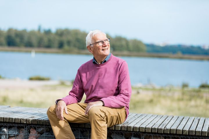 A man sits on a bench by the sea and looks up smiling.