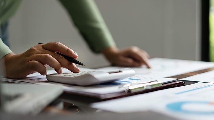 Kuvassa näkyy pöydällä olevia papereita ja kädet, jotka laskevat taskulaskimella.