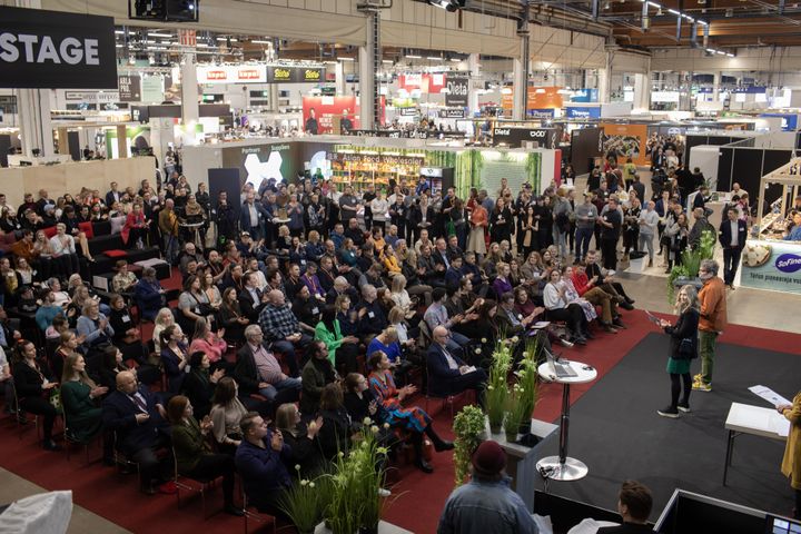 Esitys suurelle yleisölle messutapahtumassa, ympärillä useita näytteilleasettajia ja kaupallisia kylttejä.