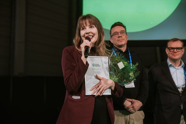Nainen puhumassa mikrofooniin, kädessään palkinto ja kukkia kahden miehen seistessä hänen takanaan.