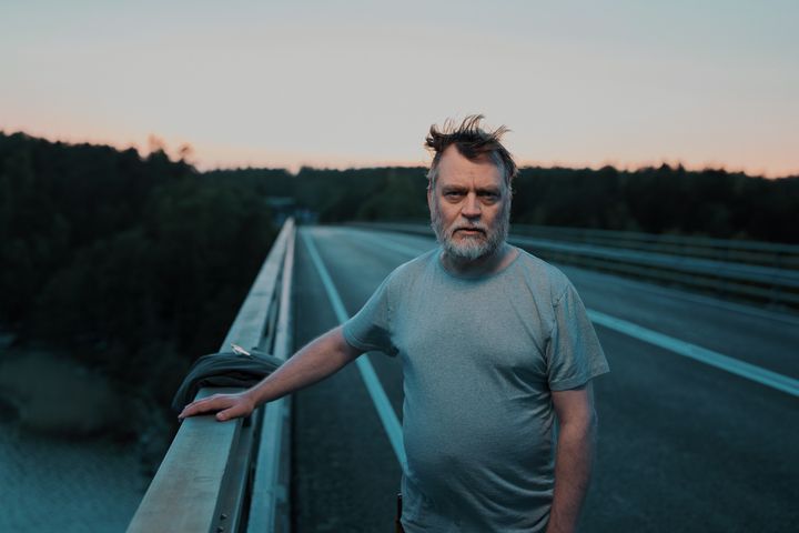 Harmaaseen t-paitaan pukeutunut keski-ikäinen mies seisoo sillalla ja katsoo vakavana kameraan. Takana näkyy metsämaisemaa ja maantie.