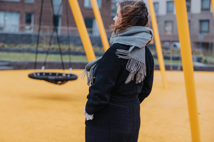 Henkilö seisoo keltaisella leikkikentällä kasvot kääntyneenä pois kamerasta. Päällä tumma takki ja harmaa huivi. Taustalla keinu.