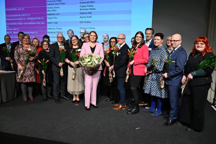 Ryhmä ihmisiä seisoo lavalla, useimmat pitävät kukkia kädessään. Keskellä on henkilö ison kukkakimpun kanssa. Taustalla on näyttö, jossa on tekstiä.