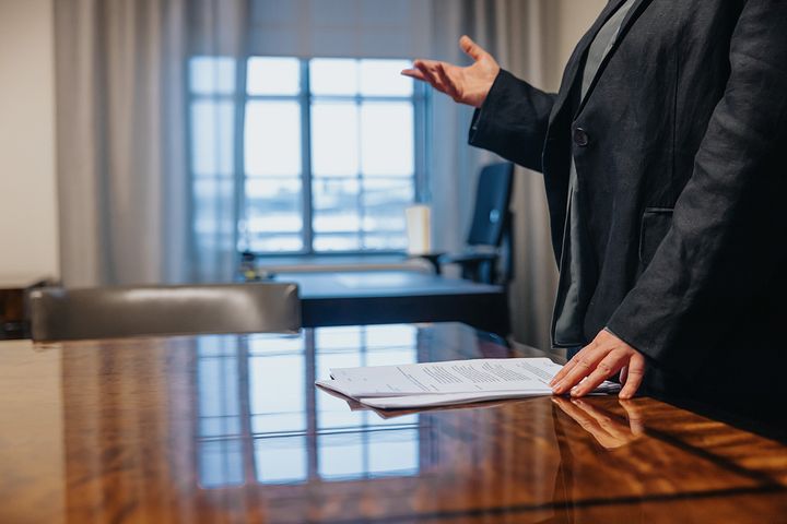 Henkilö seisoo kokoushuoneessa puupöydän ääressä, käsissään paperi.