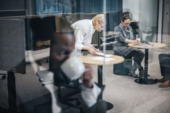 Kaksi henkilöä istuu pyöreiden pöytien ääressä toimistossa; toinen työskentelee keskittyneesti ja toinen katsoo kännykkää. Edustalla henkilö juo kahvia.