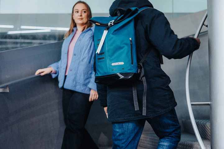 Kaksi ihmistä portaikossa, toinen kulkee ylös selkä kameraan päin, sininen reppu selässään, toinen on menossa alas ja katsoo eteenpäin.