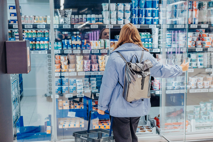 Nainen ostoskorin kanssa seisoo jogurttihyllyn edessä supermarketissa.