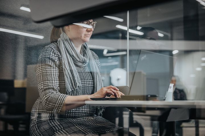 Nainen työskentelee kannettavalla tietokoneella lasiseinäisessä toimistossa.