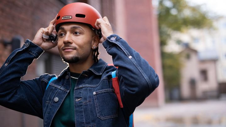 Nuori aikuinen laittaa pyöräilykypärää päähän punatiilisen rakennuksen edessä.