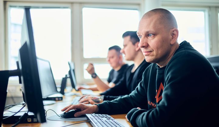 Man vid dator på kontor med två kollegor i bakgrunden.