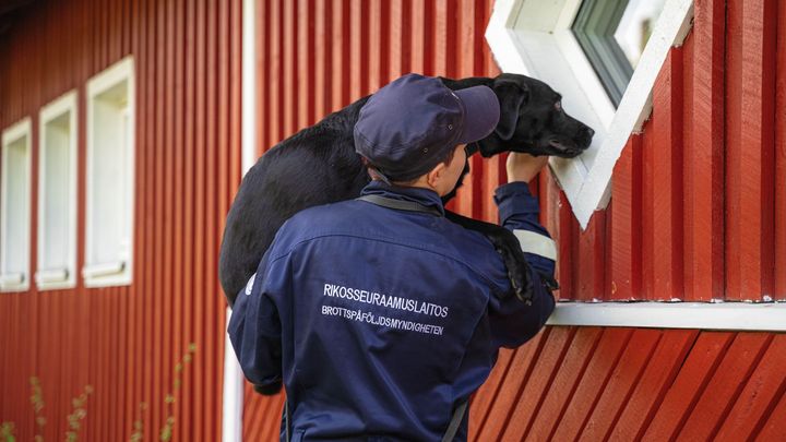 Fängelsehund och hundförare i tävlingssituation vid inspektion av en röd byggnad.