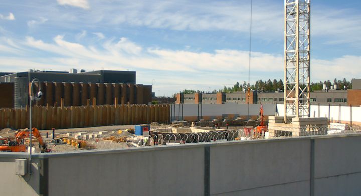 Stor och inhägnad byggarbetsplats. Grävmaskin, betongpelare och byggmaterial på plats.