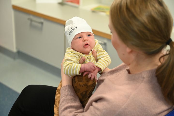 Nainen pitelee sylissään pientä sylivauvaa, jolla on päässään valkoinen Muumi-pipo.