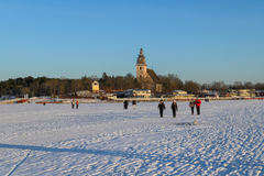 Ihmisiä käveleämässä merenjäällä Naantalinaukolla, taustalla vierasvenesataman rantaa sekä Naantalin luostarikirkko, aurinkoinen talvipäivä.