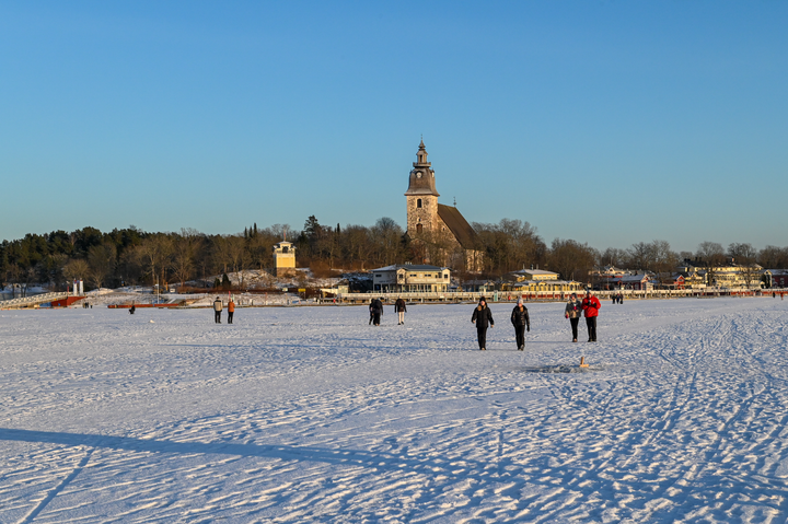 Ihmisiä käveleämässä merenjäällä Naantalinaukolla, taustalla vierasvenesataman rantaa sekä Naantalin luostarikirkko, aurinkoinen talvipäivä.
