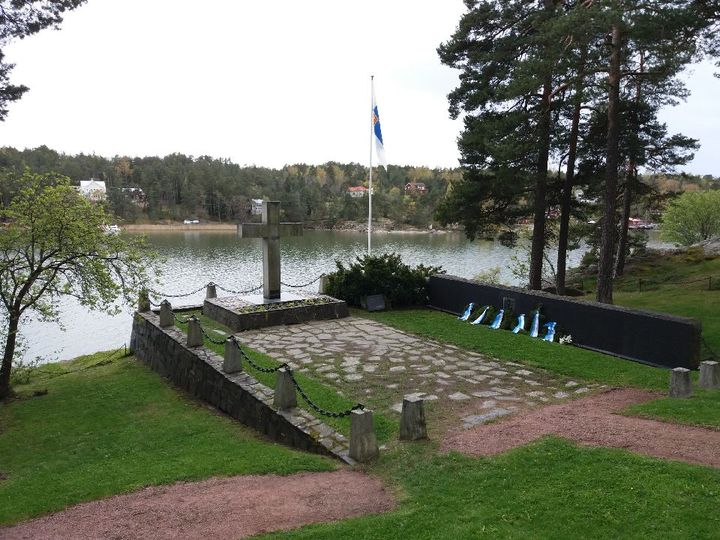 Kivinen muistomerkki, lipputanko ja muistoseinä meren rannalla.