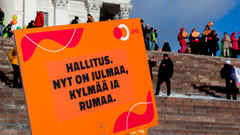 Oranssi kyltti, jossa teksti "Hallitus. Nyt on julmaa, kylmää ja rumaa". Taustalla Helsingin tuomiokirkko ja portaat, aurinkoinen talvisää.
