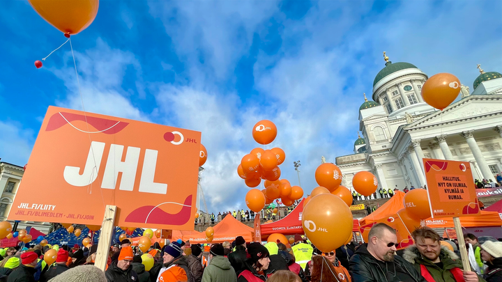 Oranssi lakkokyltti, jossa valkoinen teksti JHL, taustalla aurinkoinen Senaatintori, vaaleita pilviä taivaalla, taustalla Helsingin Tuomiokirkko.