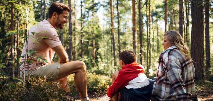 Mies-, nais- ja lapsioletettu metsässä istumassa.