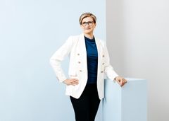 Nainen valkoisessa bleiserissä katsoo kameraan.