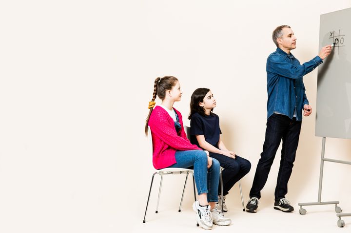 Mies kirjoittaa taululle samalla, kun kaksi istuvaa lasta seuraa häntä.