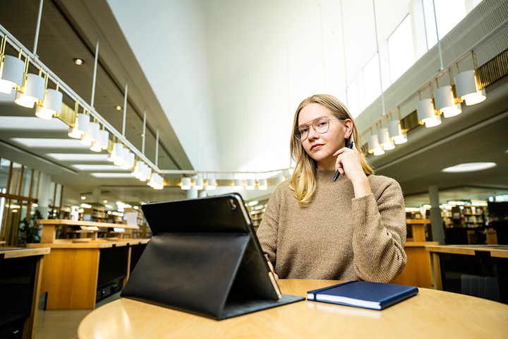 Nuori nainen kirjastossa pöydän ääressä, tabletti ja muistikirja mukanaan