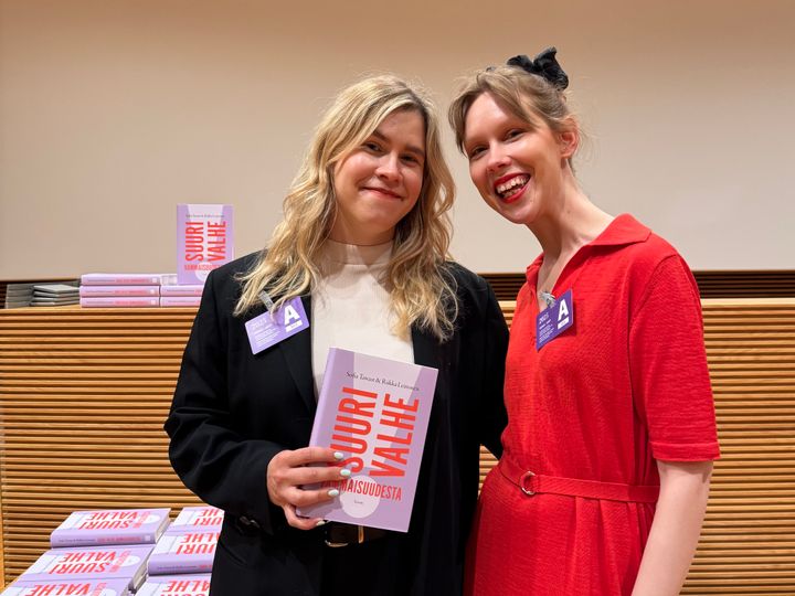 Kaksi hymyilevää henkilöä poseeraa kameralle kirjan kanssa. Kirjalla on vaaleanpunainen kansi.