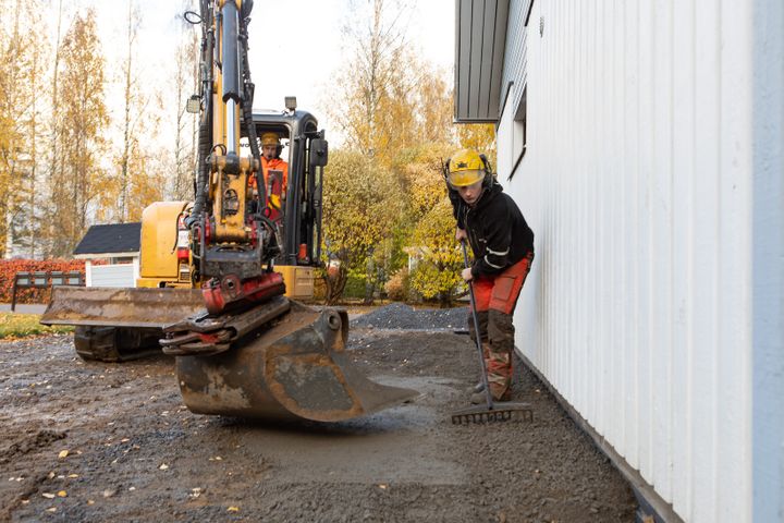 Kaksi henkilöä työskentelee ulkona kaivinkoneen kanssa talon seinän vieressä tehdessään salaojaremonttia.
