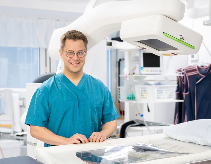 A person in a blue medical uniform stands next to medical equipment in a clinical setting.
