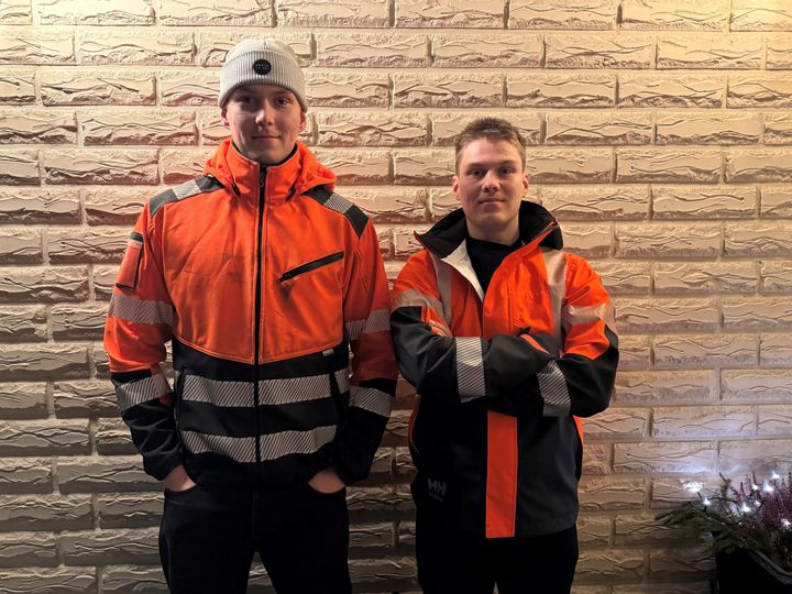 Kaksi nuorta miestä seisoo punaisissa ja mustissa ulkotakeissa vaaleaa tiiliseinää vasten.