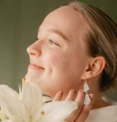 Kuvassa on henkilö, joka hymyilee ja koskettaa korvaa, johon on kiinnitetty suuret vaaleat korvakorut. Kuvassa näkyy myös kukka.