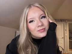 Nainen, jolla on pitkät vaaleat hiukset, nojaa käteensä ja katsoo kameraan hymyillen. Hänellä on musta paita ja vaalea huone taustalla.