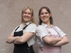 Kaksi nuorta henkilöä seisoo vierekkäin hymyillen, kädet ristissä rinnalla.